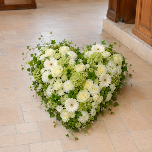 A heart-shaped floral arrangement of white and light green flowers with green ivy, placed on a beige tiled floor in a church or hall setting.