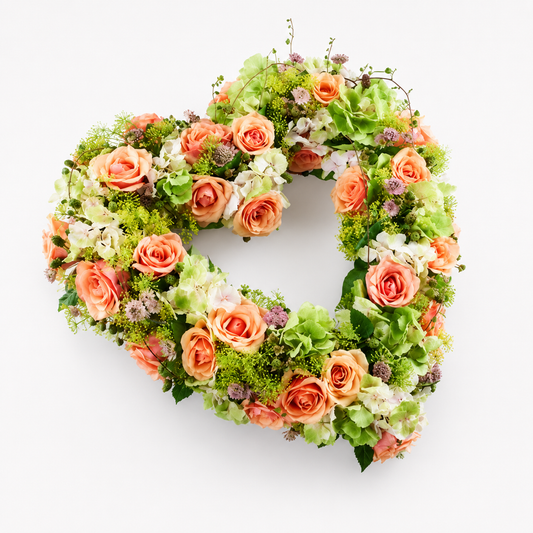 A heart-shaped floral arrangement featuring peach and coral roses, green hydrangeas, and textured green foliage, photographed from directly above on a clean white background.