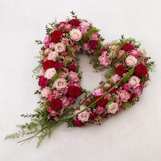 A heart-shaped floral arrangement featuring red and pink roses, light pink lilies, and green foliage, photographed from above on a plain white background.