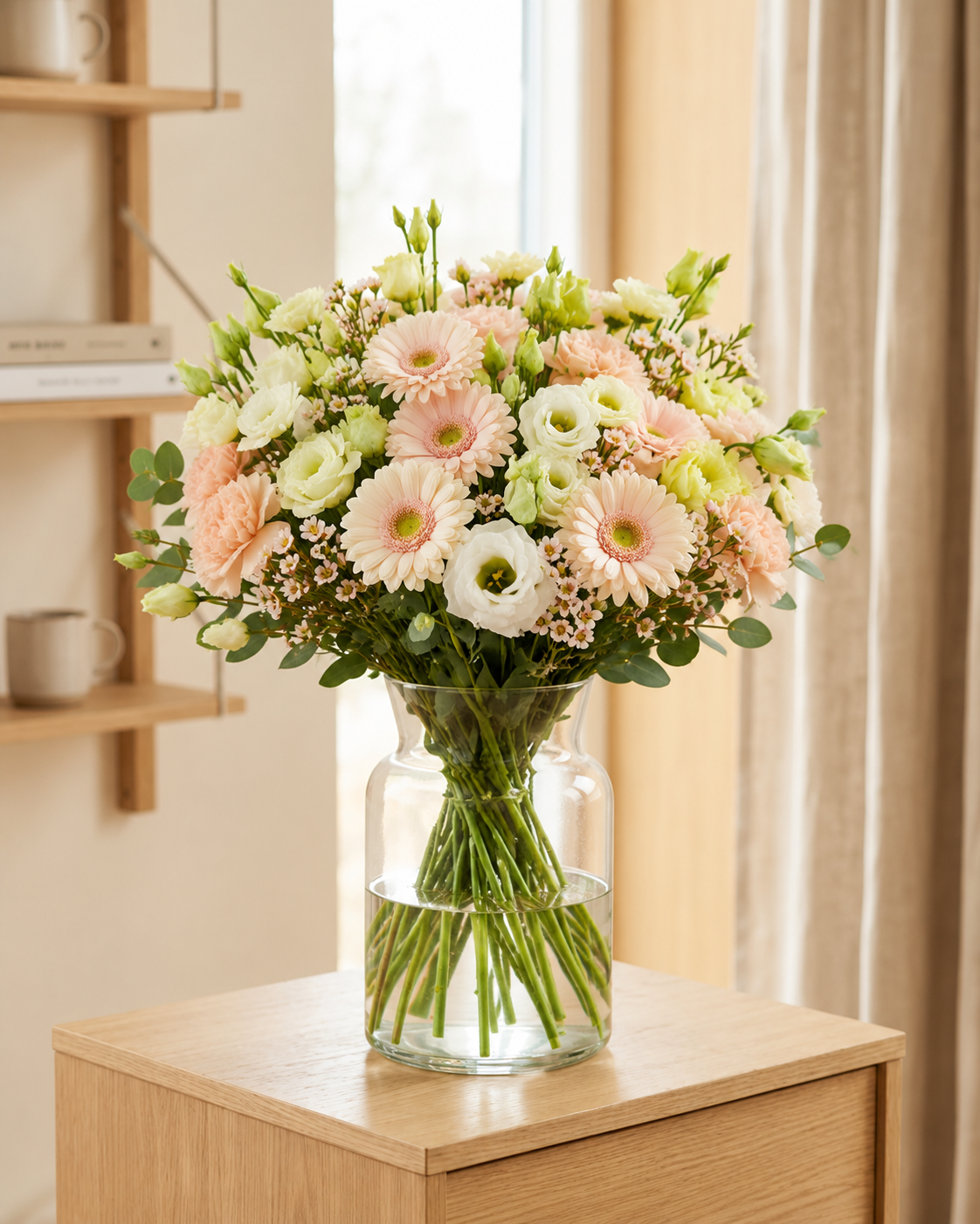 A large, vibrant floral bouquet featuring pink and white gerbera daisies, blush peonies, and green eucalyptus stems, arranged in a clear glass vase on a light wood side table.