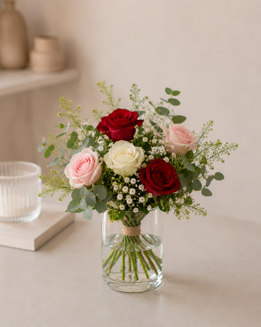 A small floral bouquet in a clear glass vase on a tabletop, featuring red and pale pink roses, a cream rose, green eucalyptus leaves, and baby's breath, with a softly blurred indoor background.