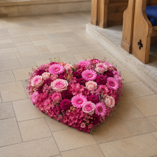 A heart-shaped floral arrangement of pink and magenta flowers, including roses and daisies, placed on a light beige tiled floor in front of wooden pews.