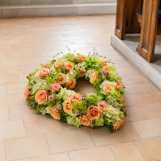 A circular floral wreath with peach roses, hydrangeas, and green foliage, placed on a beige tiled floor next to a wooden doorway.