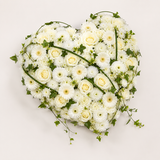 A heart-shaped floral arrangement featuring white roses, white daisies, and white carnations with green ivy vines, photographed from directly above on a light off-white background.