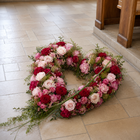 A heart-shaped floral arrangement made of pink, red, and white roses with green foliage, placed on a light beige tiled floor in a church or chapel setting.