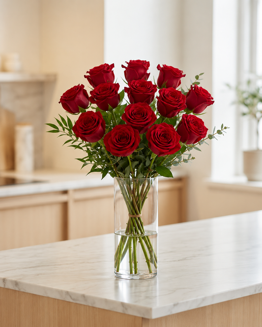 A bouquet of 12 red roses with green stems and leaves arranged in a clear glass cylinder vase, set on a light marble kitchen countertop with a blurred warm-toned kitchen background.