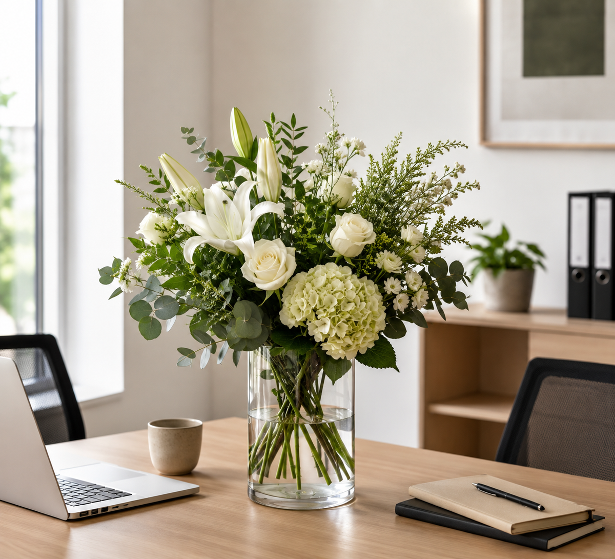 A fresh floral arrangement of white lilies, roses, hydrangeas, and green foliage in a clear glass vase, displayed on a wooden desk in a bright, modern office setting.